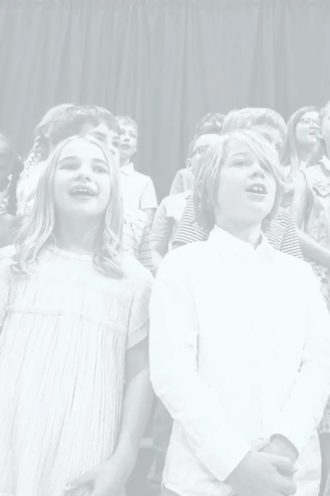 An exuberant group of children singing in a school choir, with expressions of joy and enthusiasm, against a backdrop of a dark stage curtain.