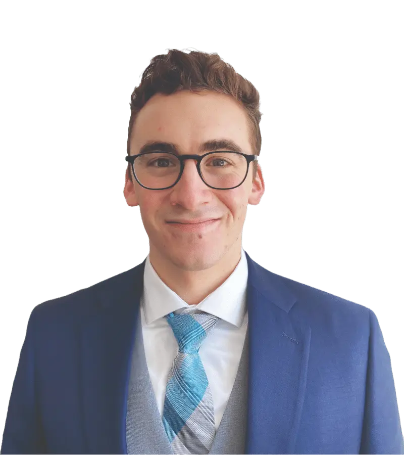 A professional headshot of a male Trinity graduate with curly hair, wearing glasses, a blue suit, and a striped tie, smiling confidently against a soft white background.