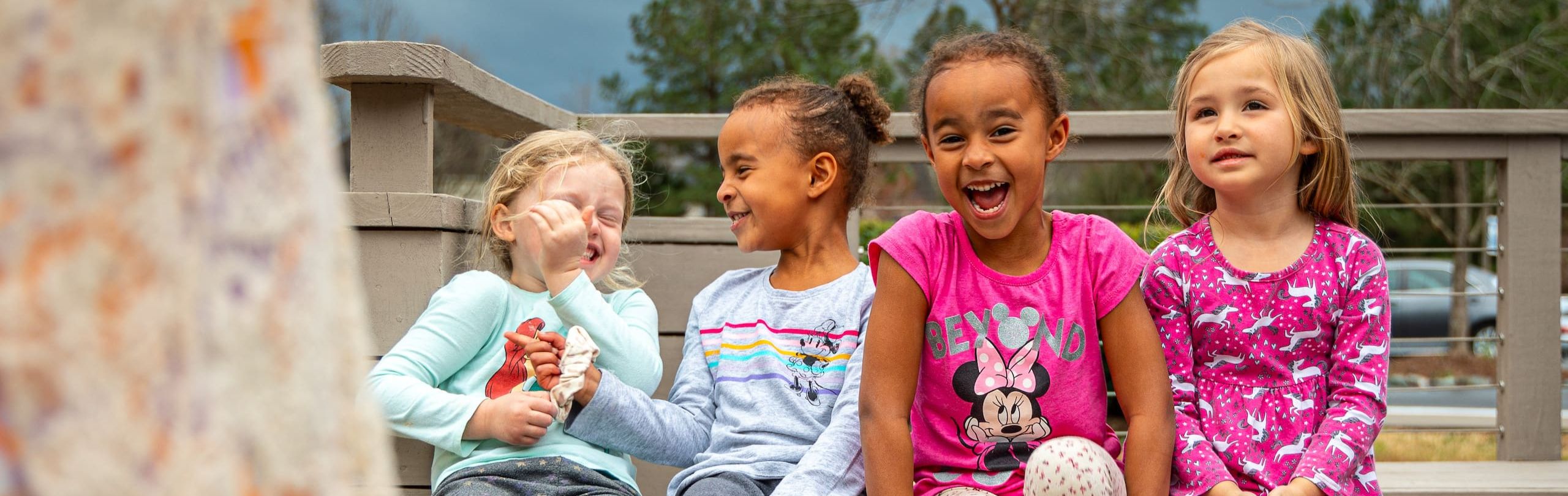 Joyful young students in vibrant attire laughing together on an outdoor bench at Trinity School, exemplifying the school's nurturing after-care program and community spirit.