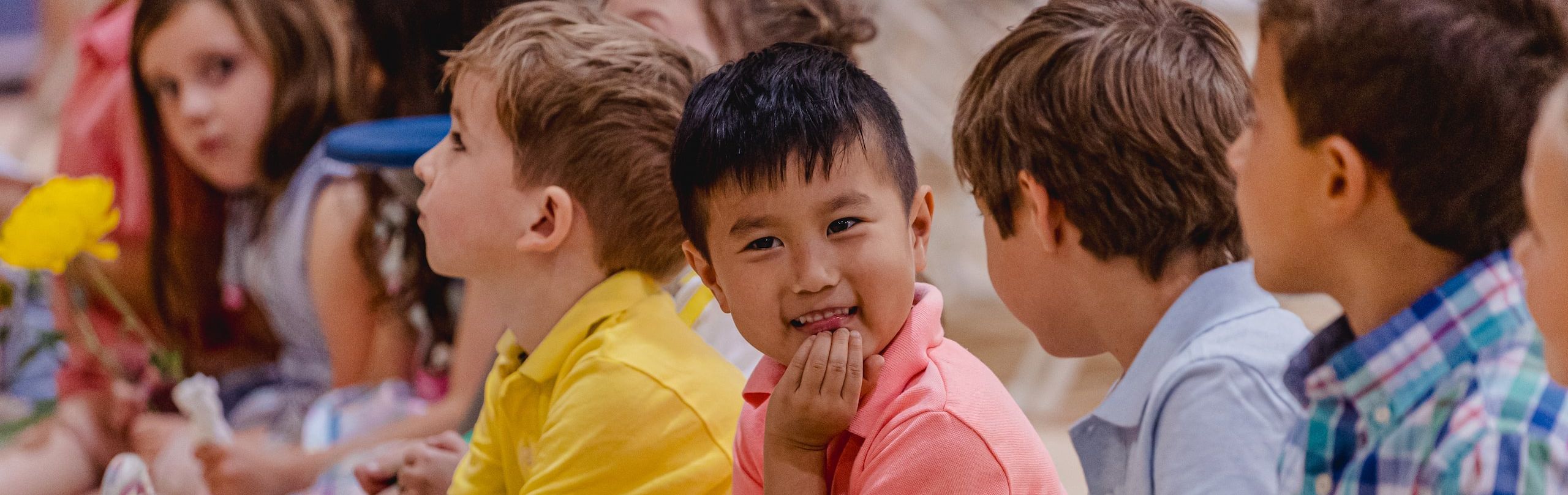 A group of attentive children sitting on the floor, one holding a yellow flower, with focus on a boy smiling at the camera.