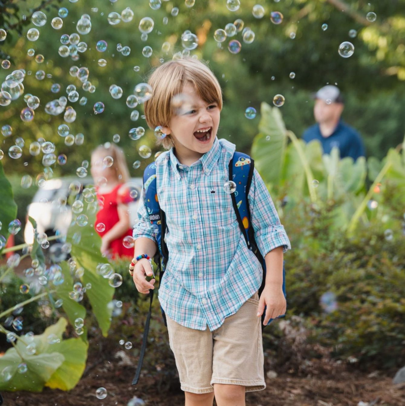 A young boy with a backpack, laughing joyously amidst floating soap bubbles with blurred figures in the background.
