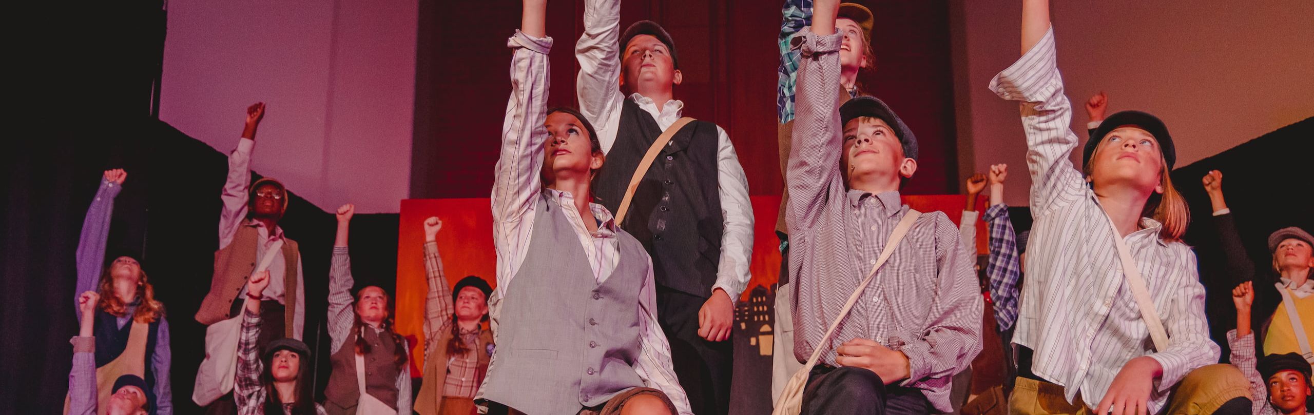 A dynamic group of middle school students, dressed in period costumes, raise their fists in unity during a spirited theater performance on stage, showcasing their acting talents.