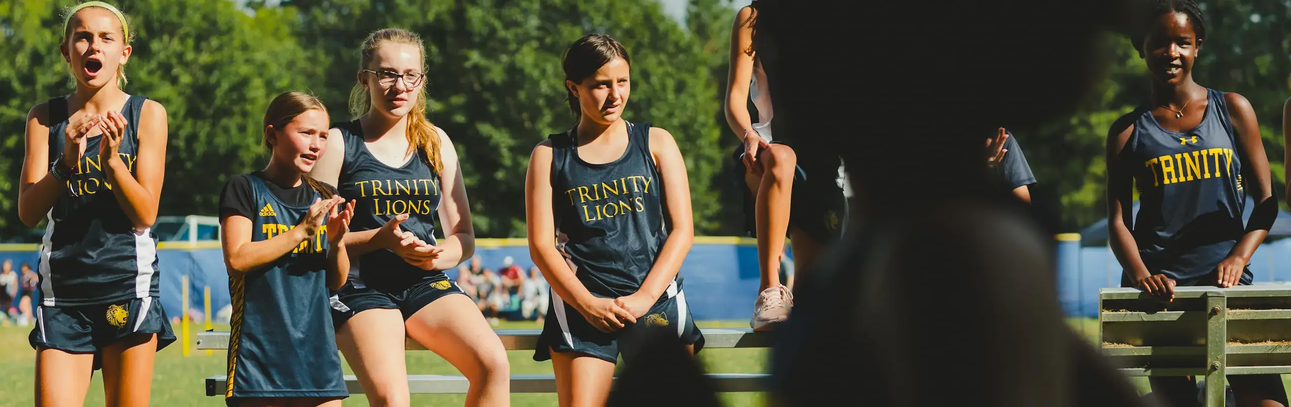 A team of young female cross country athletes in Trinity Lions uniforms enthusiastically cheering on a teammate from the sidelines on a sunny day.