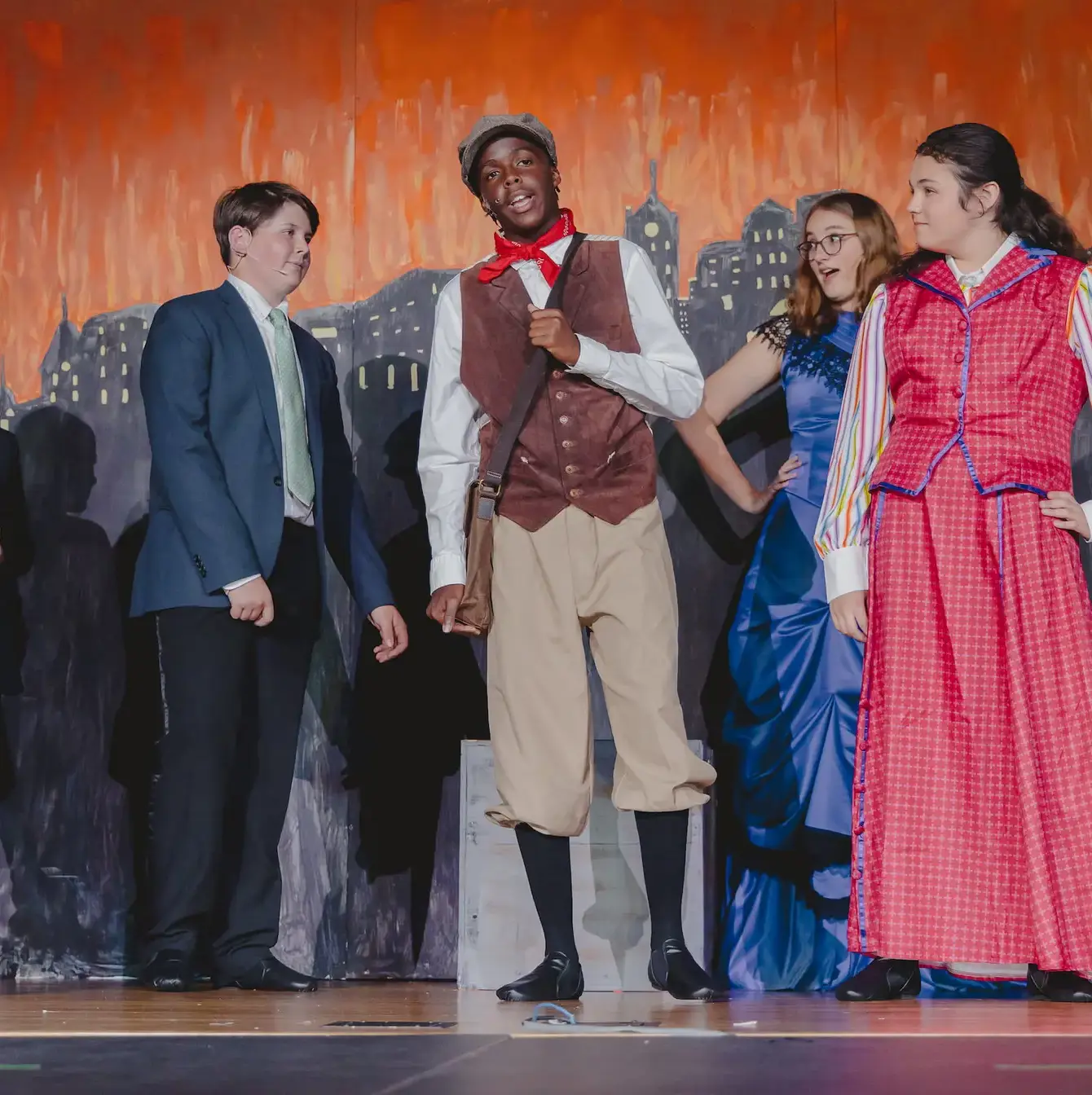 A lively scene from a middle school theater production with students in vibrant period costumes performing on stage, depicting a moment from the musical 'Newsies Jr'.