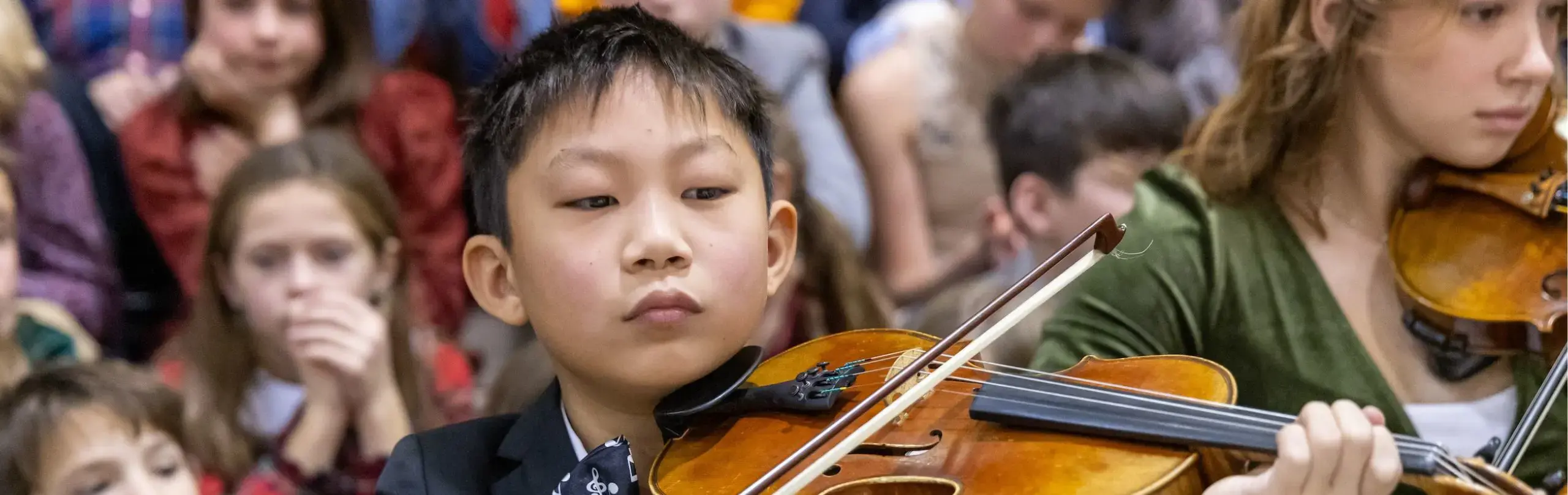 Young Asian boy playing violin focusedly at school event, surrounded by attentive classmates in audience.