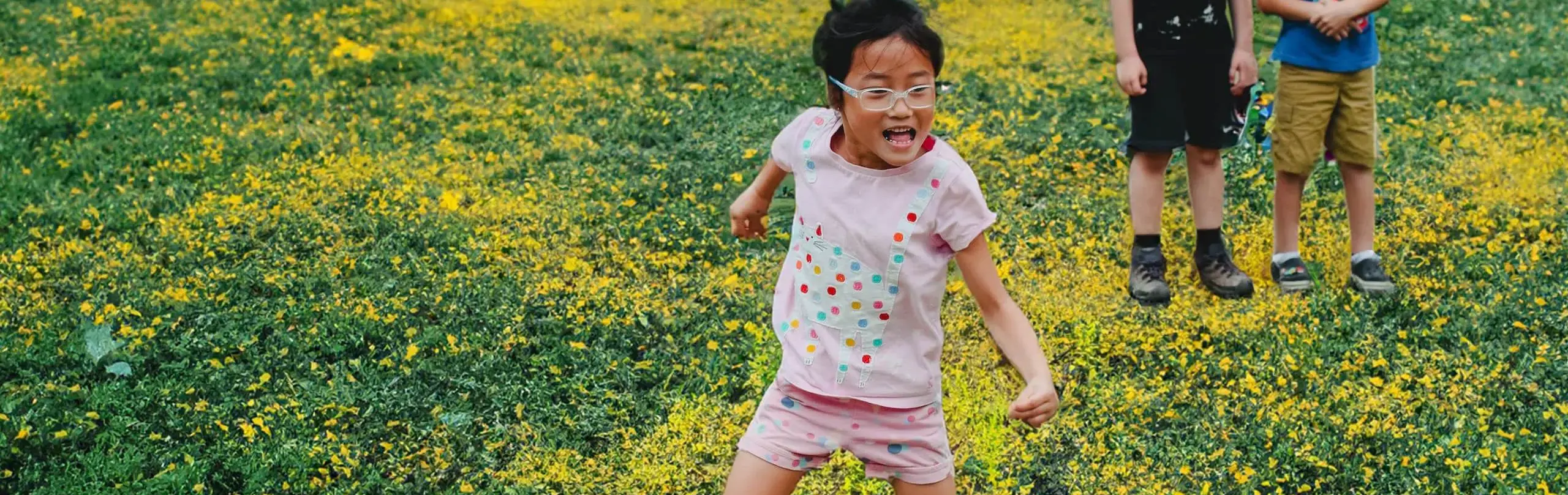A joyful girl with glasses runs through a field of yellow flowers, her excitement palpable. In the background, two boys stand watching, one with a smile, against the vibrant green and yellow backdrop.