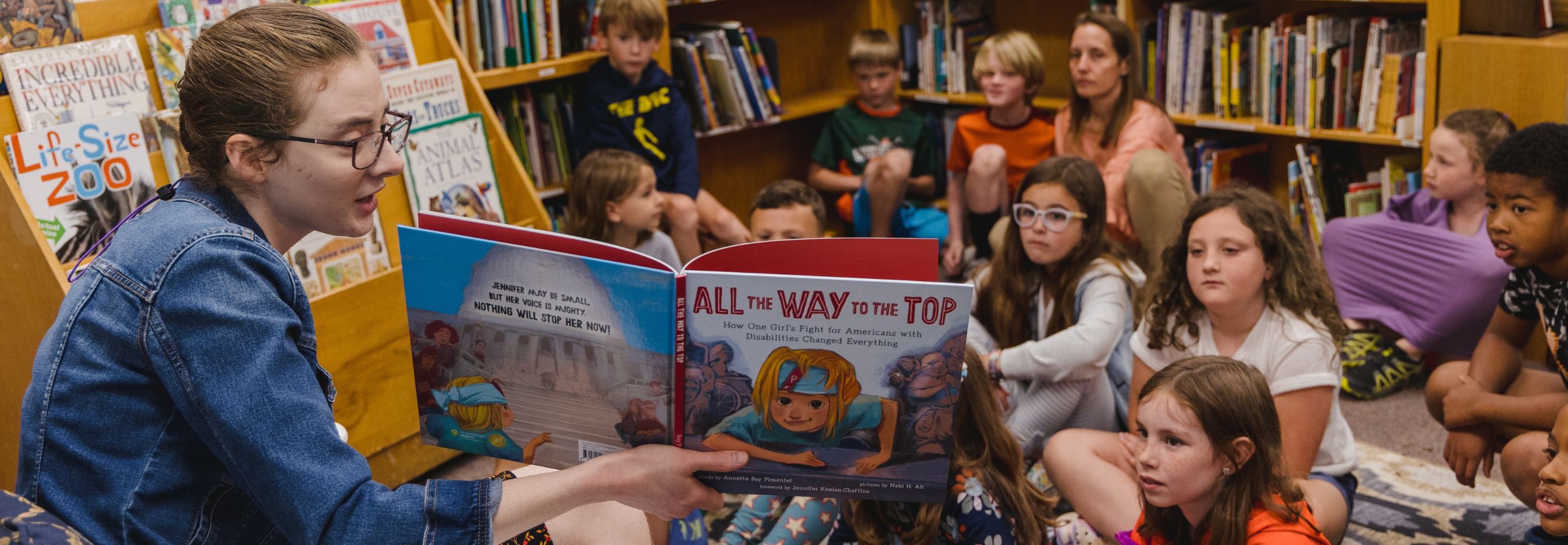 A young woman reads a picture book to a group of attentive children sitting on the floor in a cozy library setting. The bookshelves in the background are filled with colorful children's books, creatin