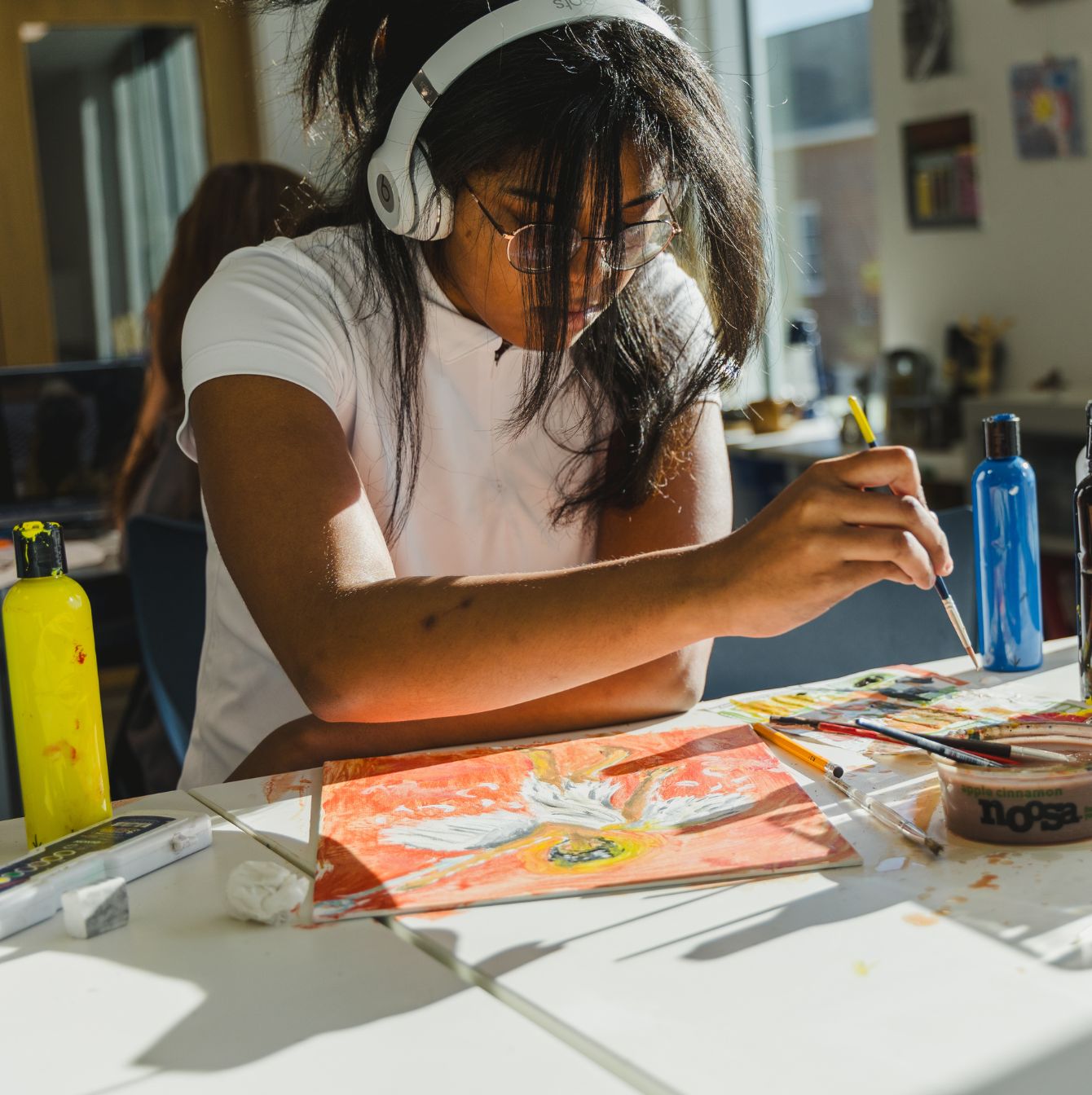 An attentive student artist is deeply focused on painting a colorful canvas in a sunlit art studio, surrounded by vibrant paint bottles and the inspiration of a bright creative space.