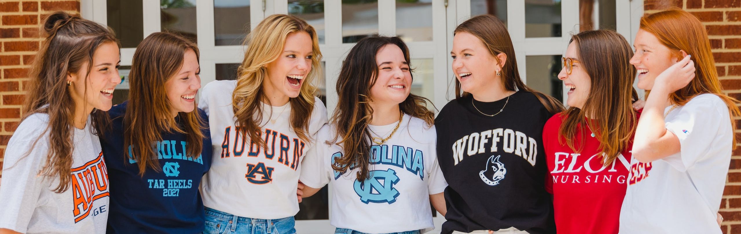 High school girls laughing together, each wearing a college t-shirt, representing the excitement and camaraderie of college decision day at school.