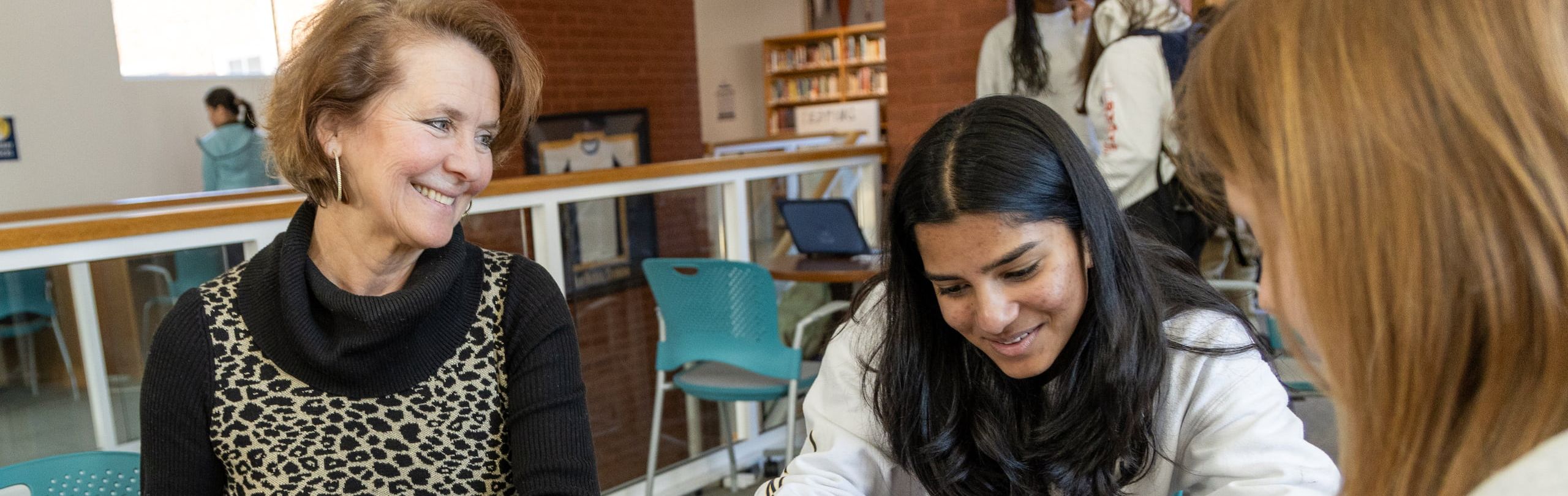 A smiling teacher engaging with a female student who is focused on writing in a notebook at a round table in a library, with other students in the background.
