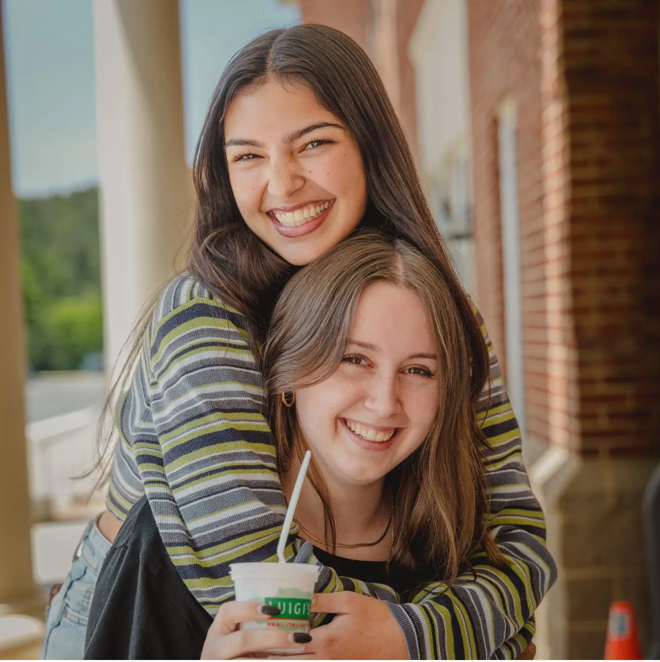 Two Trinity high school friends share a joyful hug and laughter on a sunny day; one is holding a refreshing snack, adding to the casual and happy atmosphere of their outdoor moment.
