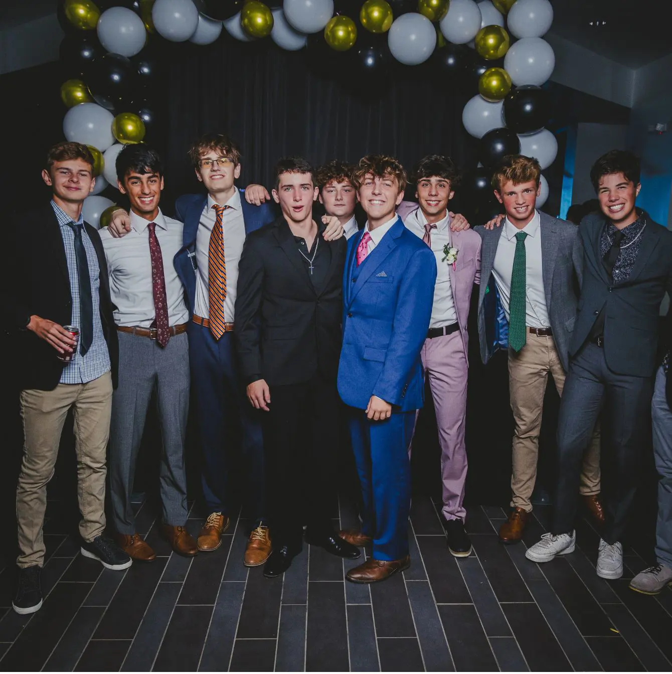 A group of Trinity high school boys in semi-formal attire posing confidently at a school event, with a backdrop of black, white, and gold balloons, radiating youthful camaraderie and celebration.