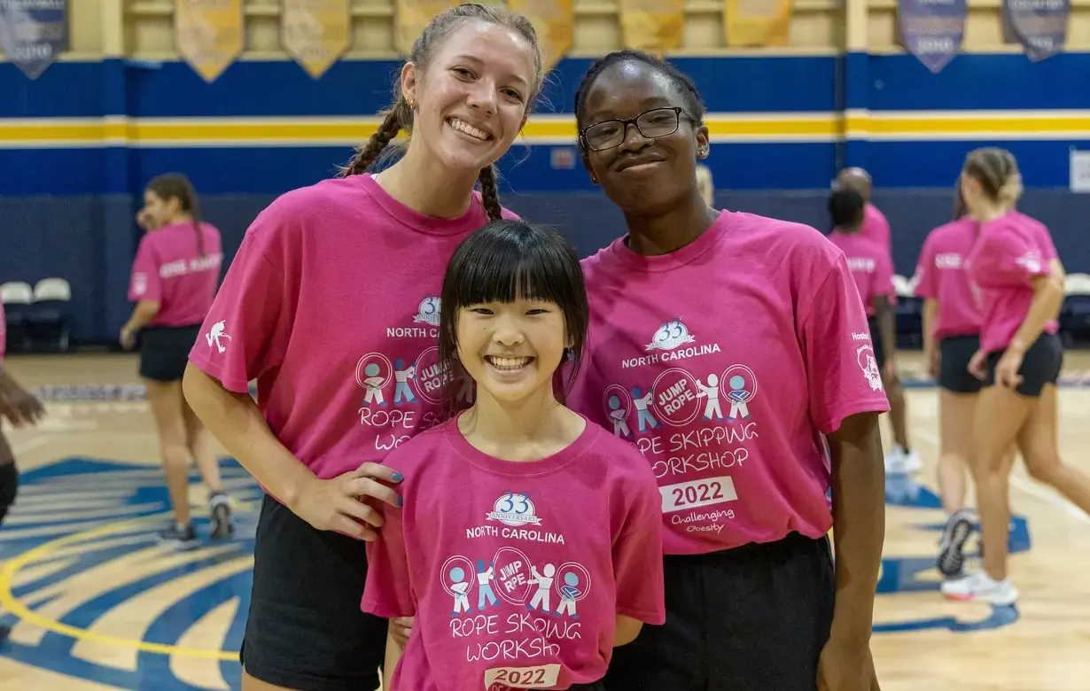 Three young Bouncing Bulldog girls in matching pink t-shirts with a rope skipping workshop logo, smiling together in a gymnasium with sports banners in the background.On Page Image