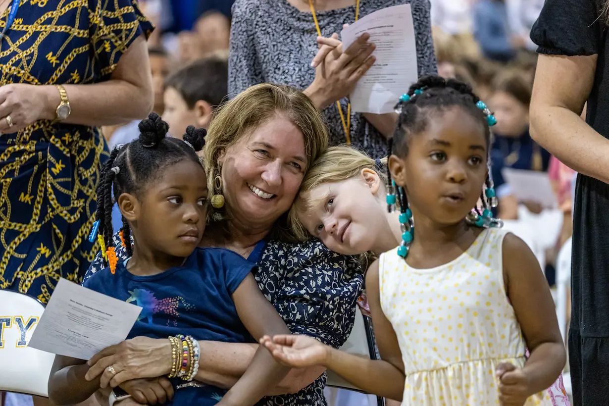 A joyful Trinity Teacher hugging two young children in a chapel service with other attendees in the background.