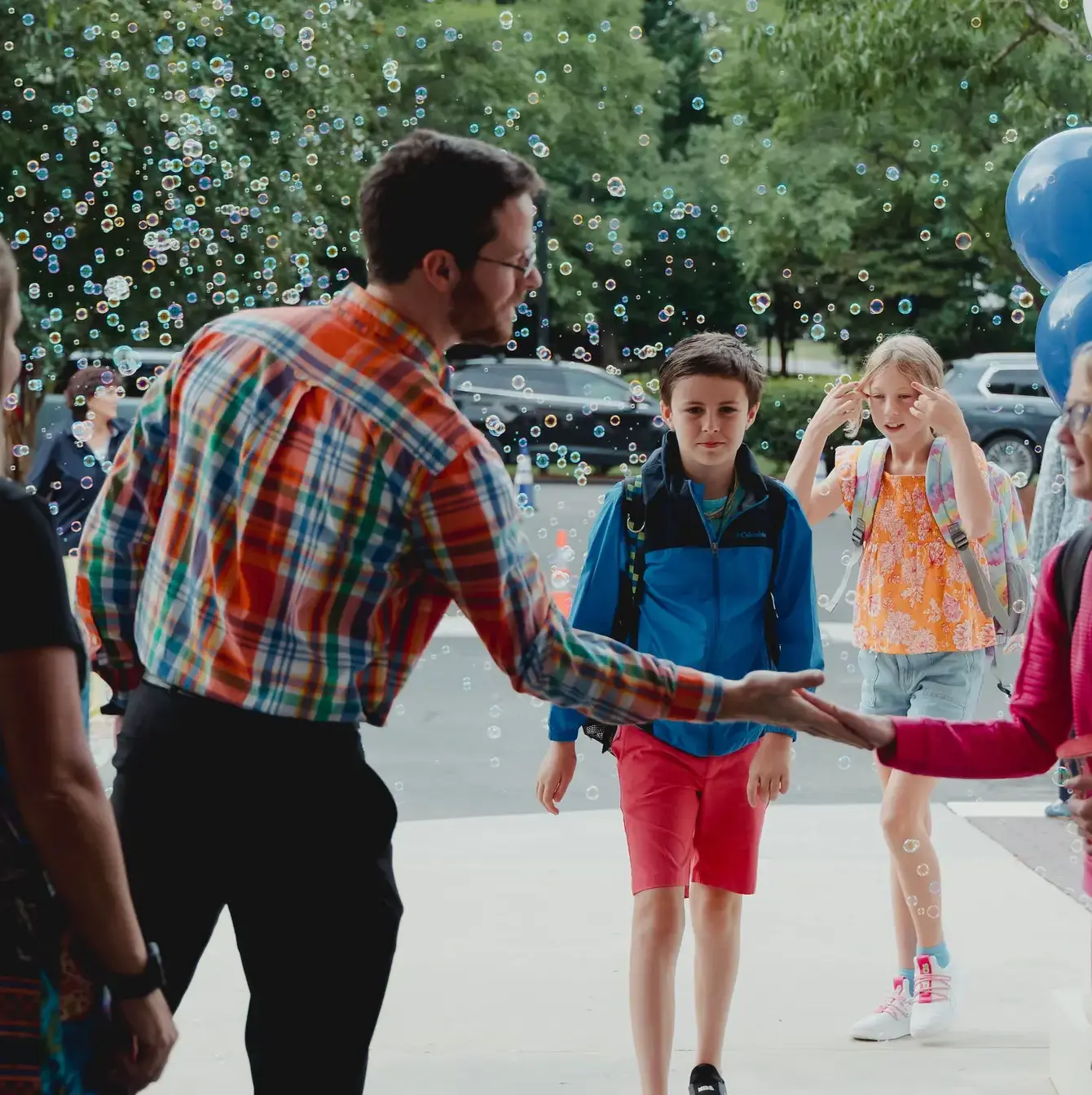 A teacher in a colorful plaid shirt greets students with a handshake at the school entrance, surrounded by a festive atmosphere with floating bubbles and balloons in the background.