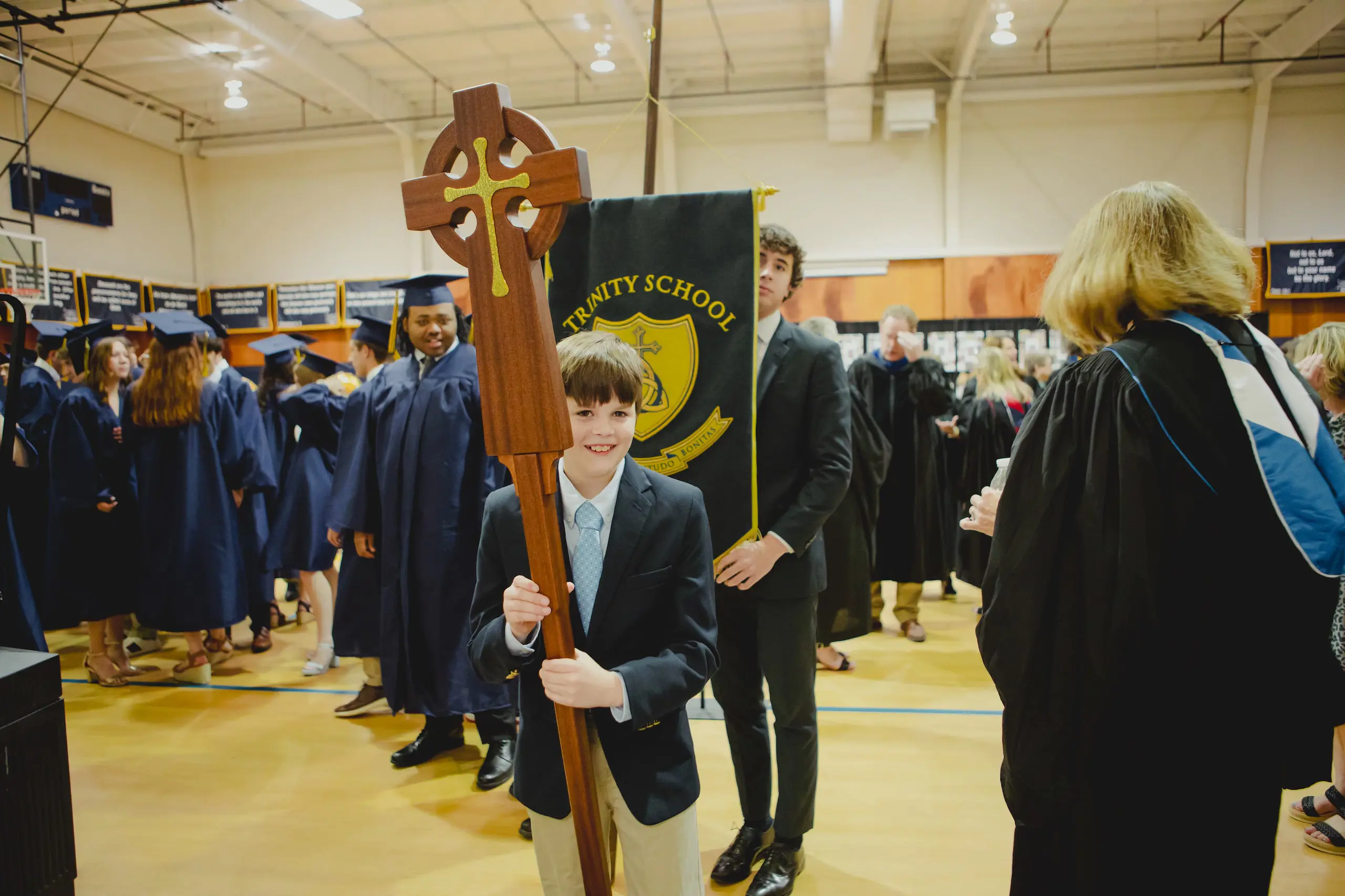 A young Trinity student smiling as he holds a cross during a procession, with high school seniors in graduation gowns following behind in a gymnasium.