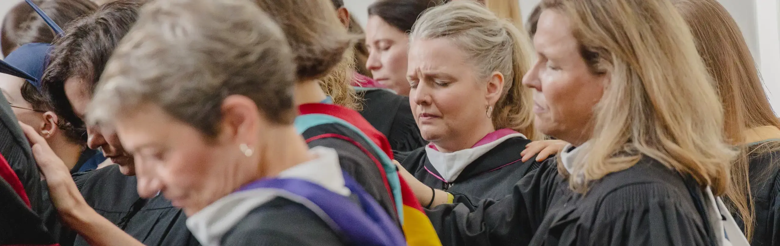 A group of female faculty members bow their heads in a solemn moment of prayer, wearing traditional black graduation gowns during a prayer ceremony for seniors about the graduate.