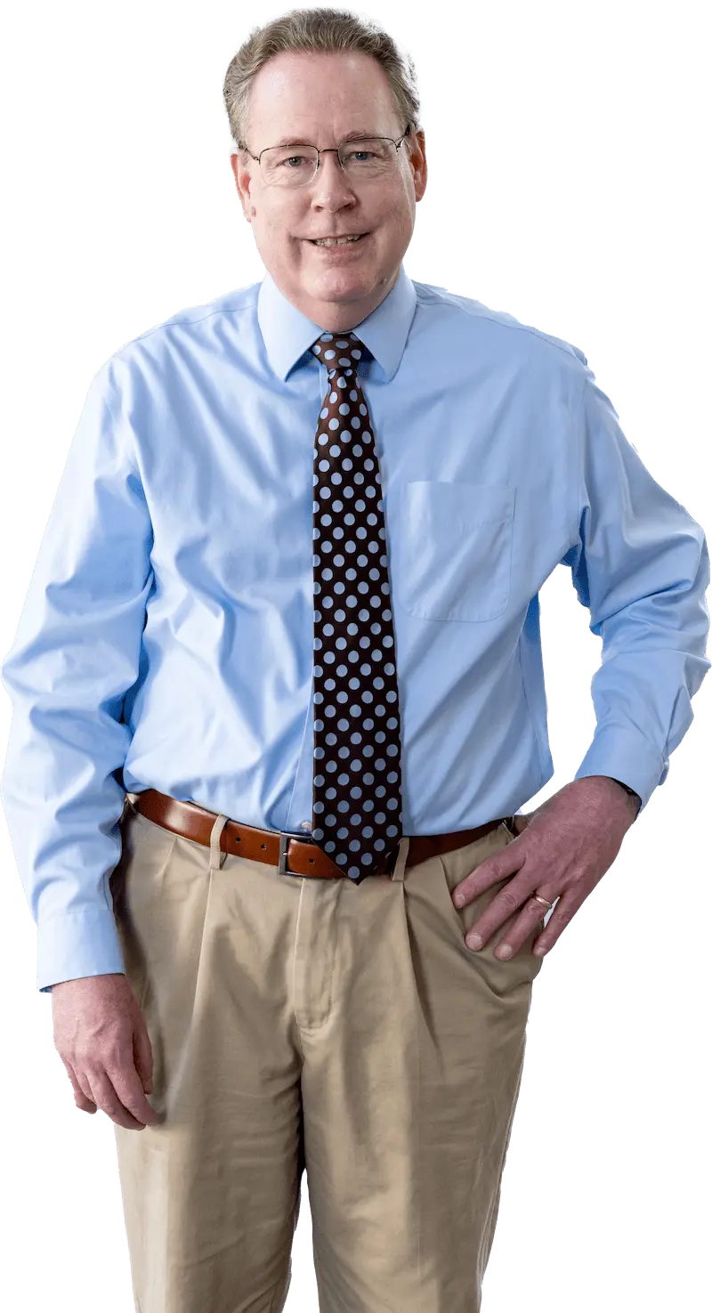 Professional portrait of a smiling middle-aged man with glasses, wearing a light blue shirt, polka dot tie, and khaki trousers, standing with hands on hips against a black background.