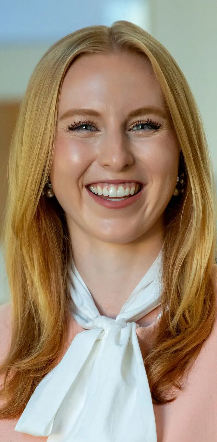 A cheerful blonde woman wearing a peach blazer and white tie-neck blouse, smiling warmly.