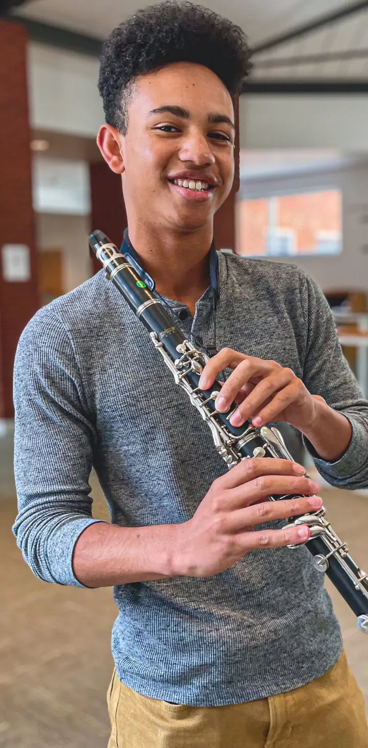 A young man with a bright smile holding a clarinet, standing in an indoor setting with modern architecture.