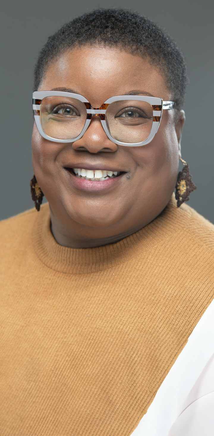 A headshot of a confident professional woman with a beaming smile, wearing large fashionable glasses and a caramel sweater, set against a neutral gray background.