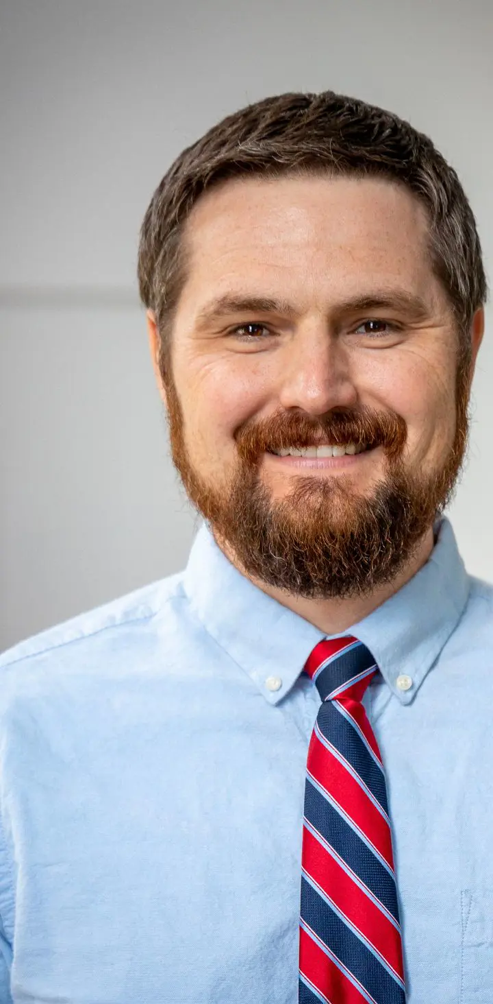 A professional headshot of a male educator with a beard, wearing a light blue shirt and a red striped tie, smiling warmly with an American flag partially visible in the background, conveying a sense o