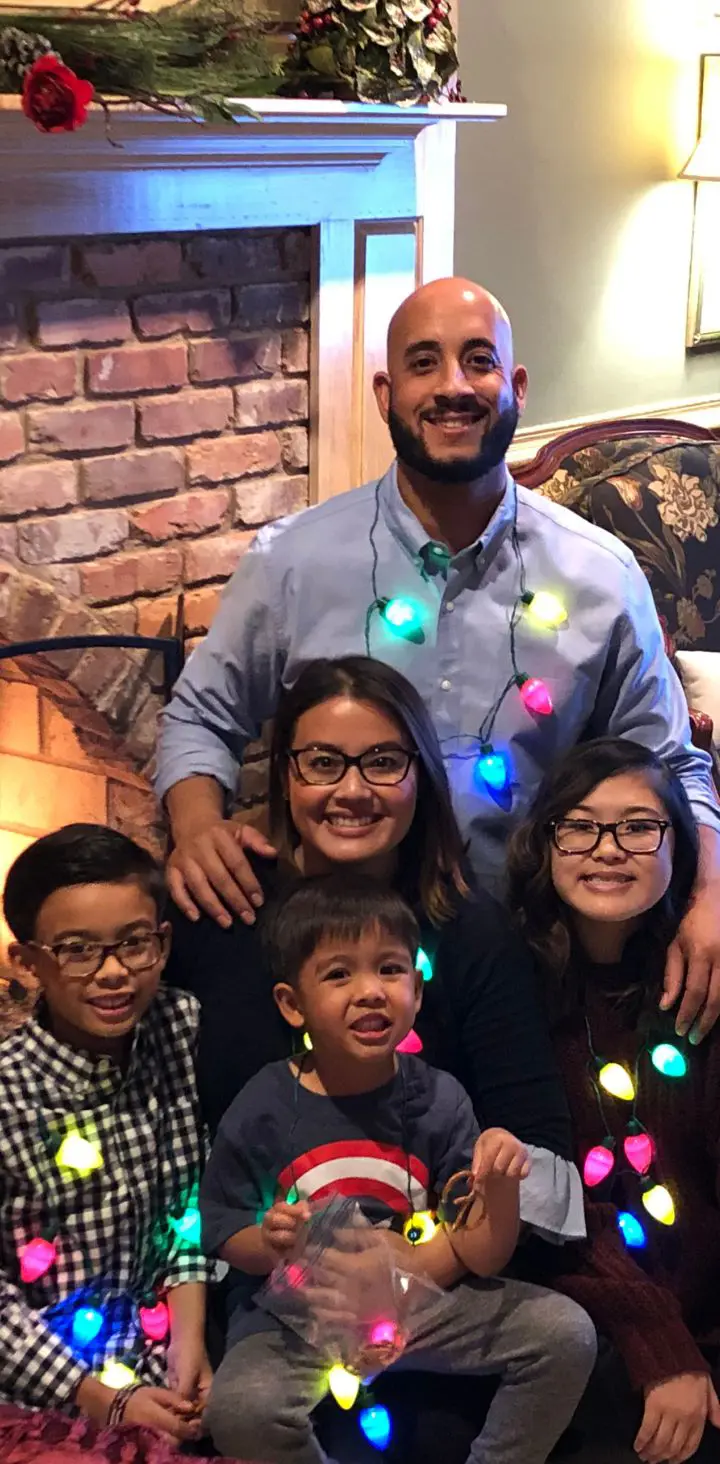 Happy family of five with three children, wearing Christmas lights as necklaces, sitting together in a living room decorated for the holidays with a lit fireplace and festive decorations.