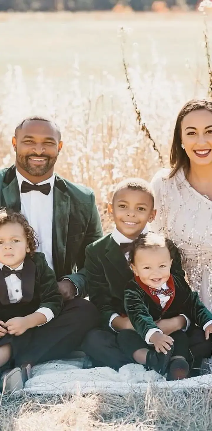 A joyful family dressed in formal attire poses for a holiday portrait outdoors, with four smiling children and a couple seated on a blanket amidst tall grasses, under the clear sky.