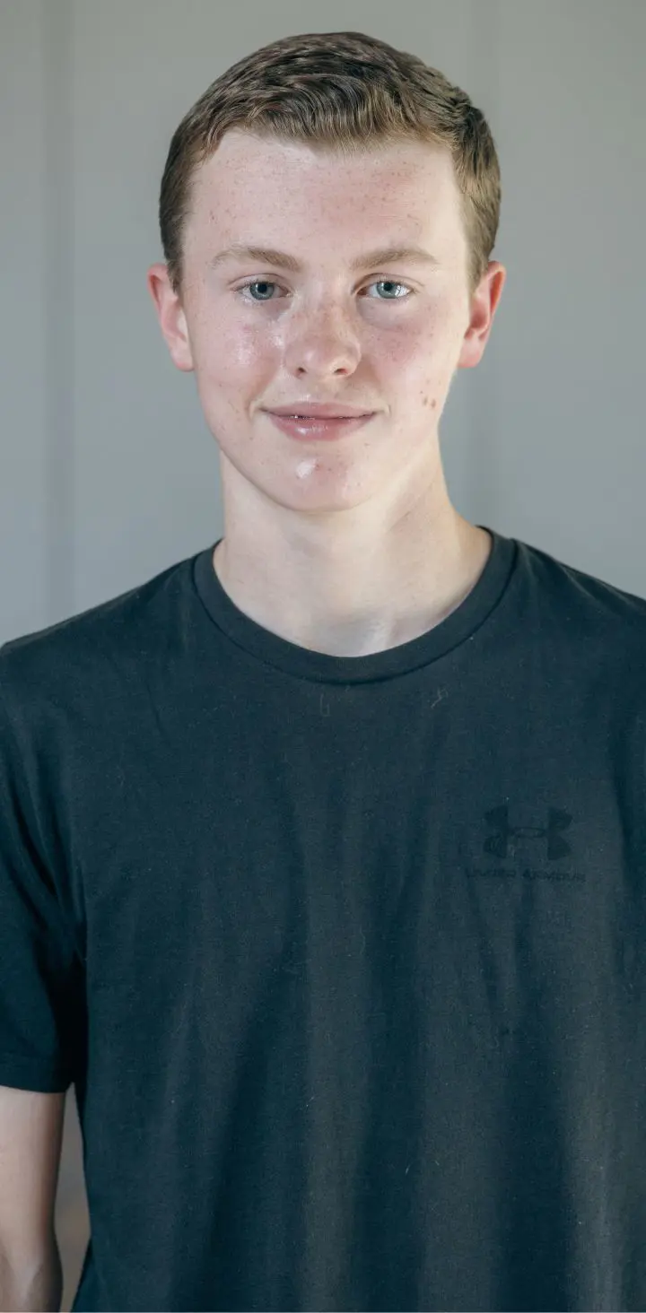 A headshot of a confident teenage male Trinity student with a slight smile, wearing a black t-shirt, standing against a neutral background with natural light highlighting his features.