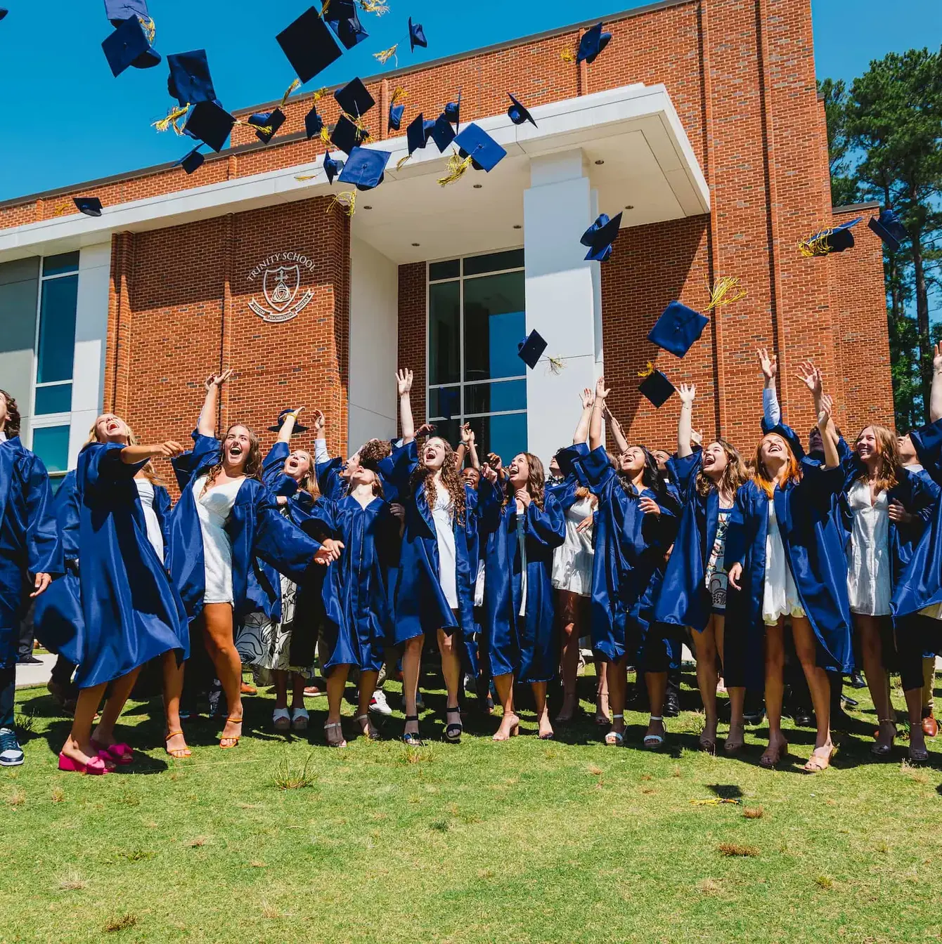 Exuberant high school graduates in blue gowns joyfully tossing their caps in the air in front of their school building.