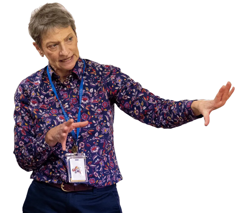 An animated female teacher explaining a concept with hand gestures in a classroom with students and educational posters in the background.