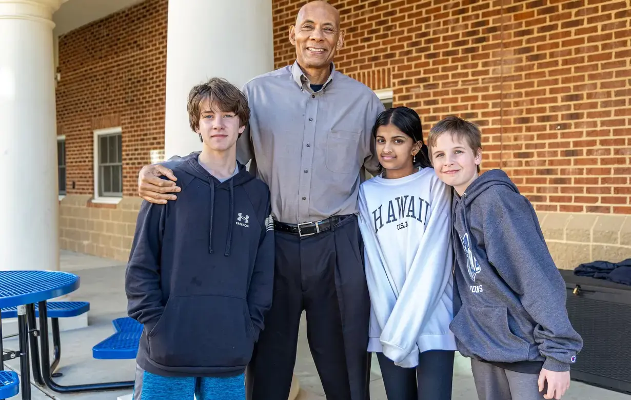 Three middle school students with diverse backgrounds smiling alongside their tall male teacher outdoors.