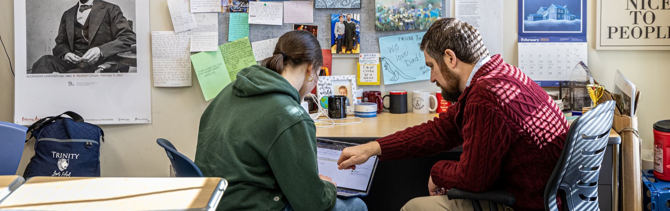 A high school student and a teacher engage in a focused mentoring session in a classroom adorned with inspirational banners and educational decor, highlighting the importance of guidance in education.