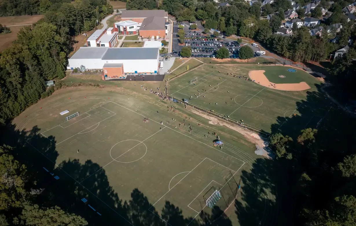 Aerial view of Trinity School campus, featuring modern buildings surrounded by lush greenery, a spacious parking lot, and sports fields in the background.