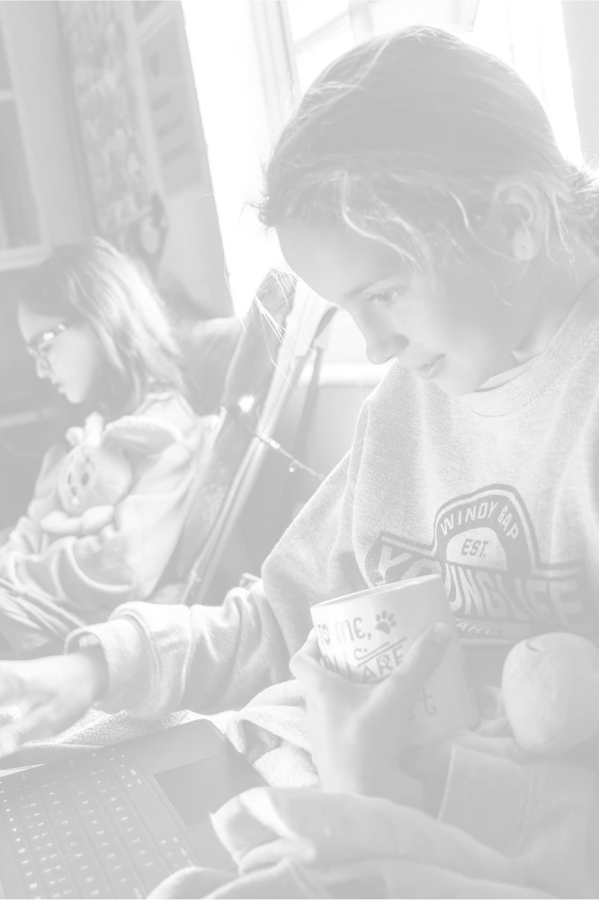 Two young girls engaged in reading and working on a laptop in a cozy room adorned with fairy lights and a bookshelf.