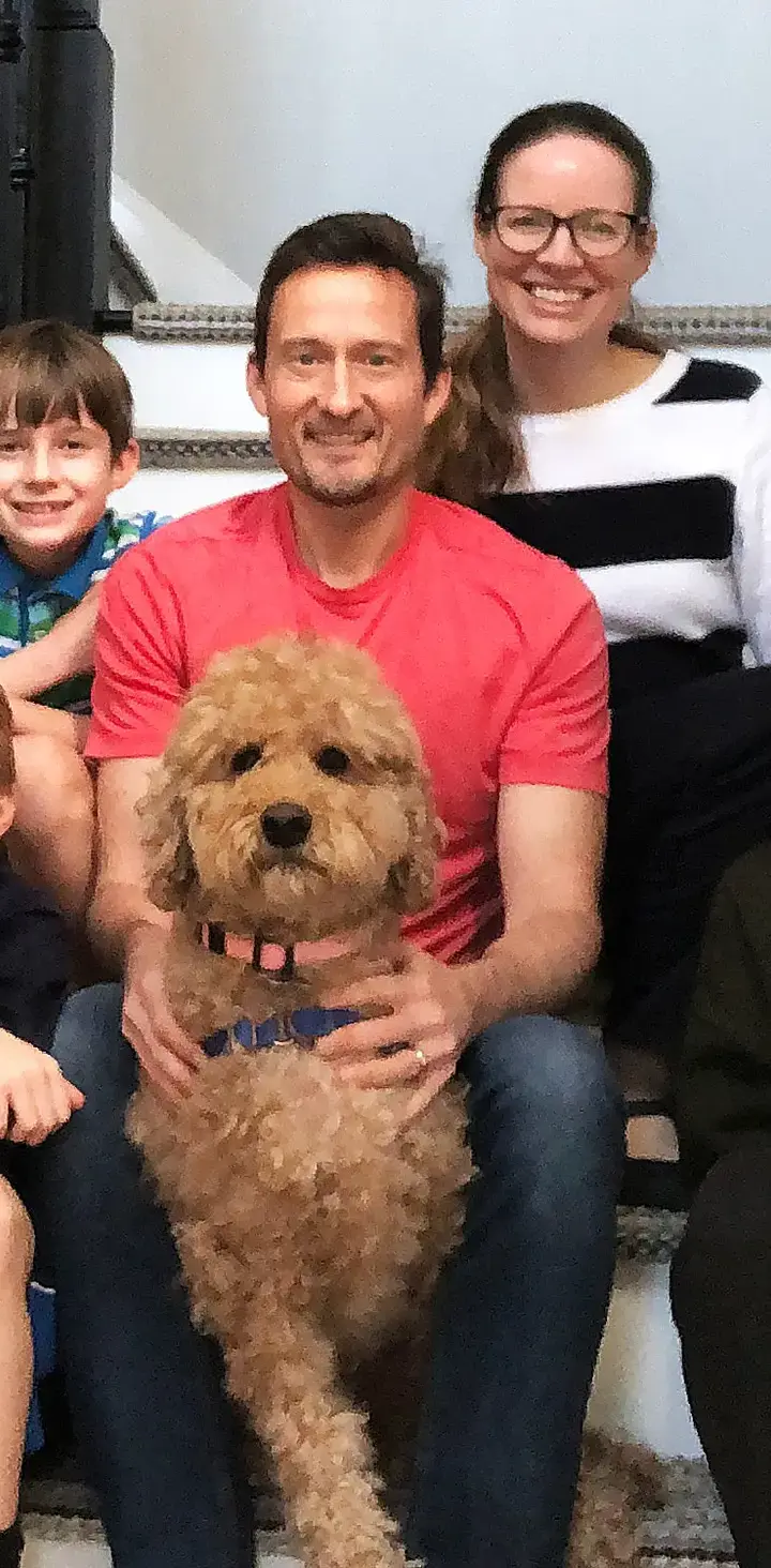 A cheerful family of five, with three children and a fluffy dog, pose for a holiday portrait on their home staircase, adorned with festive decorations and Christmas lights in the background.